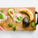 Avocado Basil Morning Smoothie. Made with avocado, basil, banana,pineapple, and coconut milk. Smoothies are on a William Sonoma charcuterie cutting board