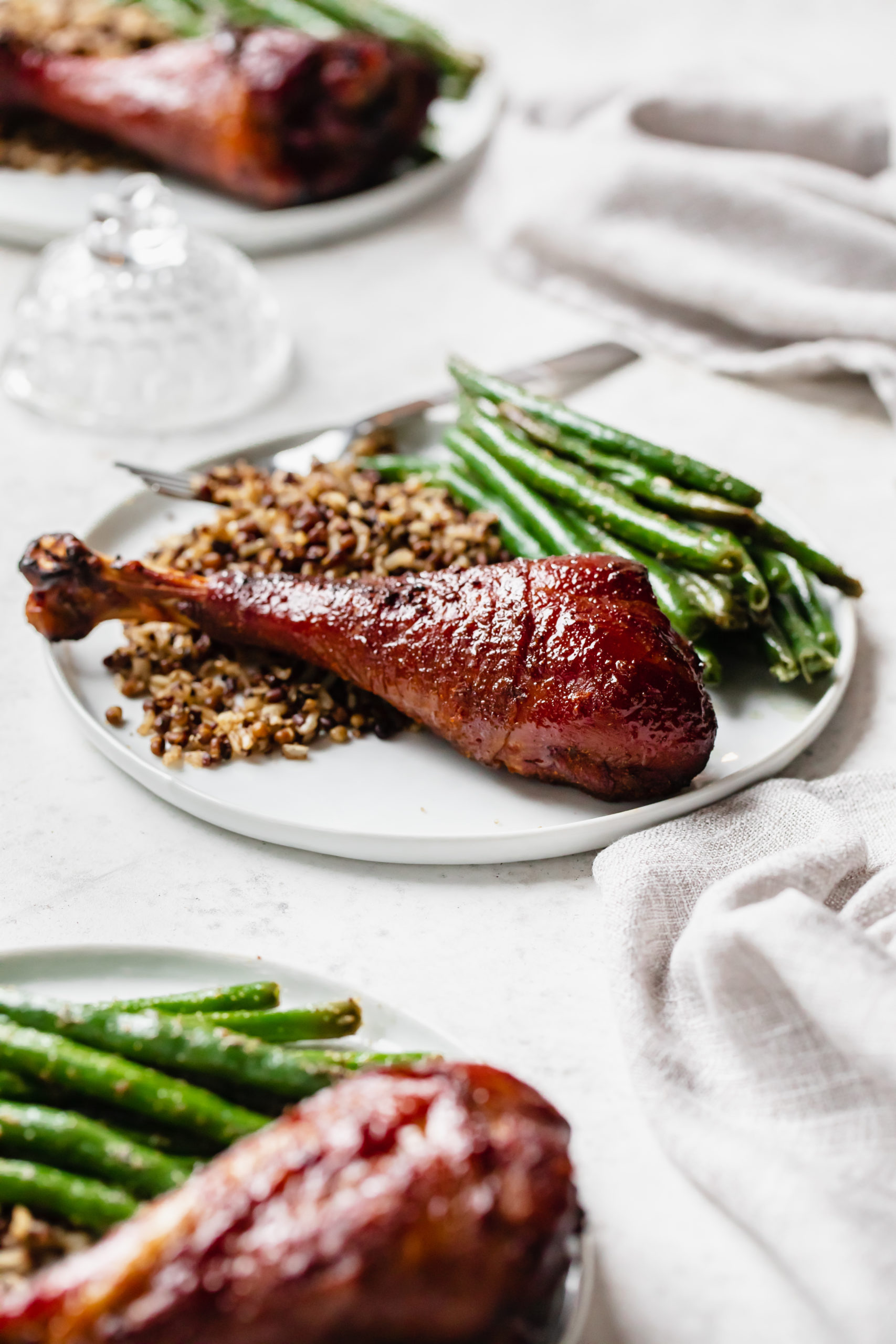 side view of turkey drumstick on circular white plate with green beans and grains. 